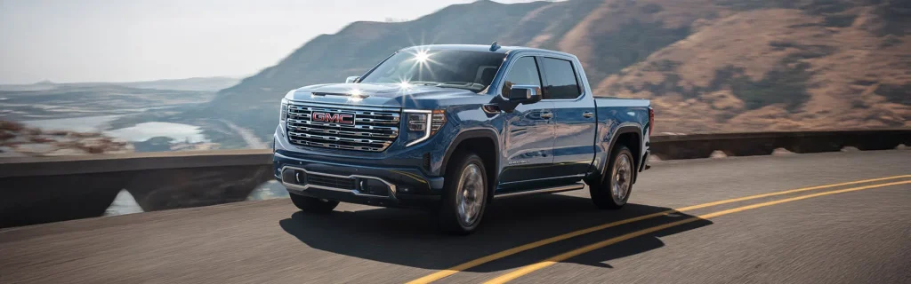Blue 2025 GMC Sierra 1500 driving along a scenic one-lane highway beside a river and mountainous terrain on a sunny day near Mangino Buick GMC.