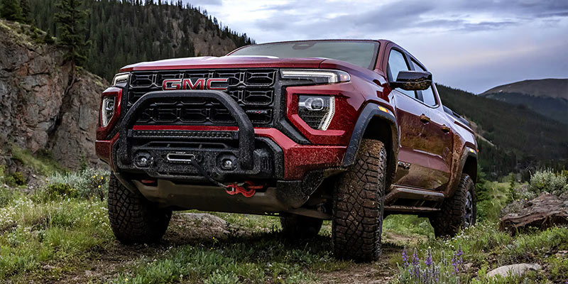 Front low-angle view of a red GMC Canyon pickup truck equipped with off-road tires and a bull bar driving through a rocky mountain trail landscape.