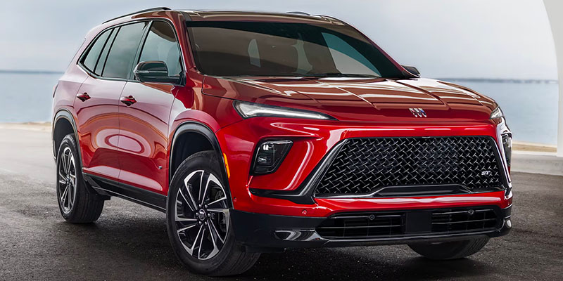 Exterior of a shiny red 2025 Buick Enclave suv as seen from the front and passenger side, parked near a body of water in New York. 