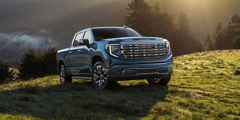 GMC Sierra 1500 parked on a grassy hillside with a forested mountain backdrop during golden hour lighting.