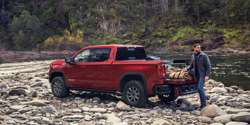 A man unloading gear from the bed of a red pickup truck on rocky terrain near a river.
