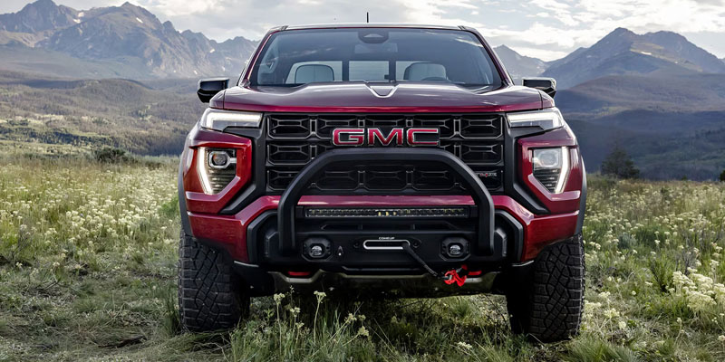 Front view of a red GMC pickup truck parked in a grassy field with mountains in the background.