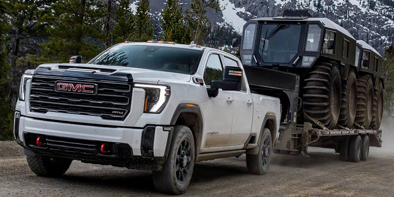 White GMC pickup truck towing a large off-road trailer through a forested landscape.