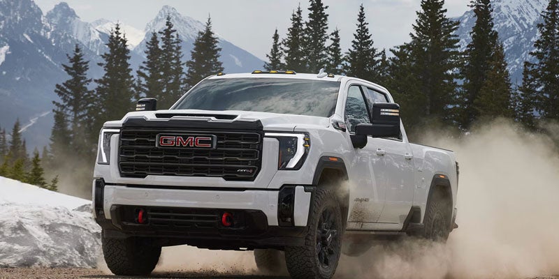 White GMC pickup truck driving through a snowy landscape with pine trees and dust rising behind it.