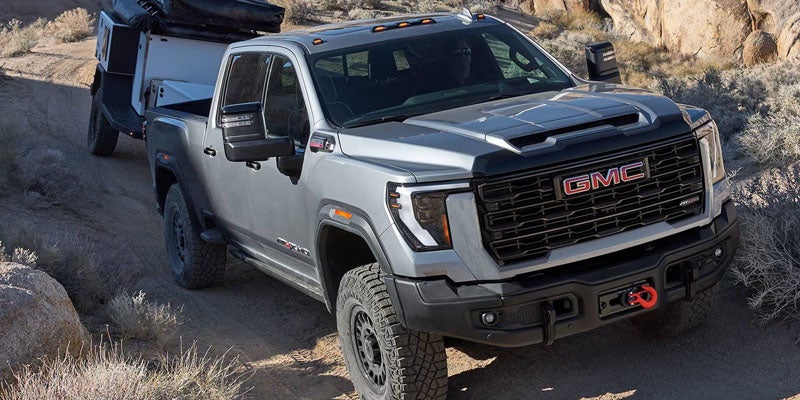 Silver GMC pickup truck driving on a rugged dirt trail with rocky terrain in the background.